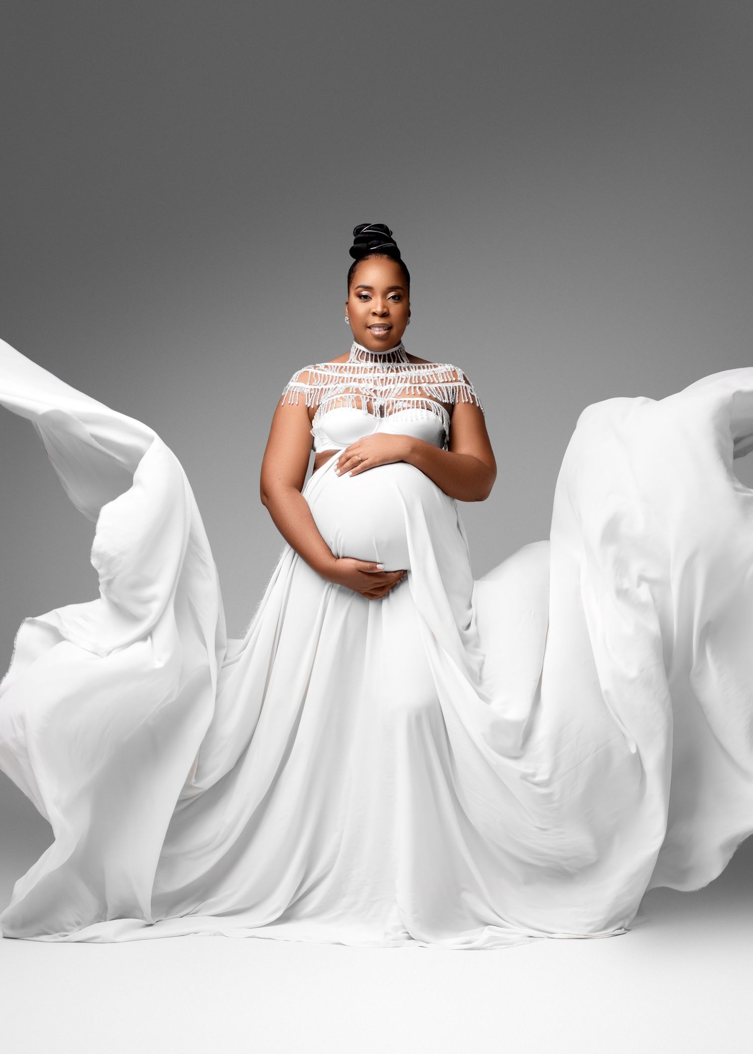 Dramatic maternity photo with flowing white fabric and lace details against a grey background.