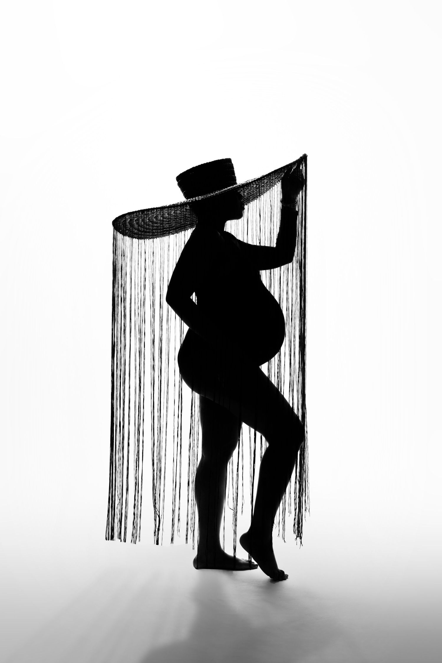 Artistic black and white silhouette of a pregnant figure behind striped curtains.