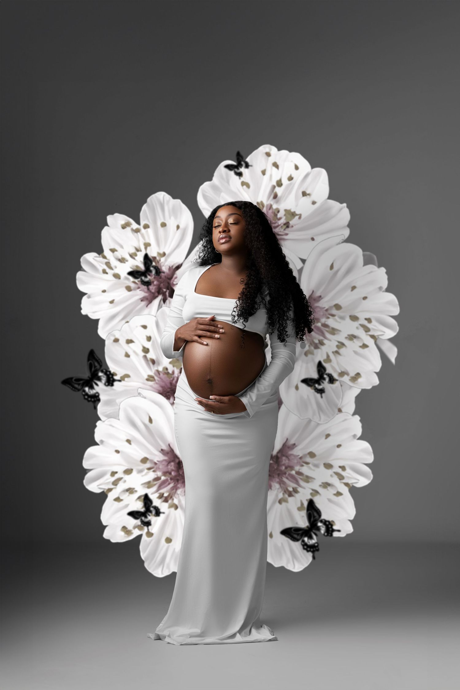 Artistic maternity photo featuring flowing white gown against large floral backdrop with black butterflies on gray background.