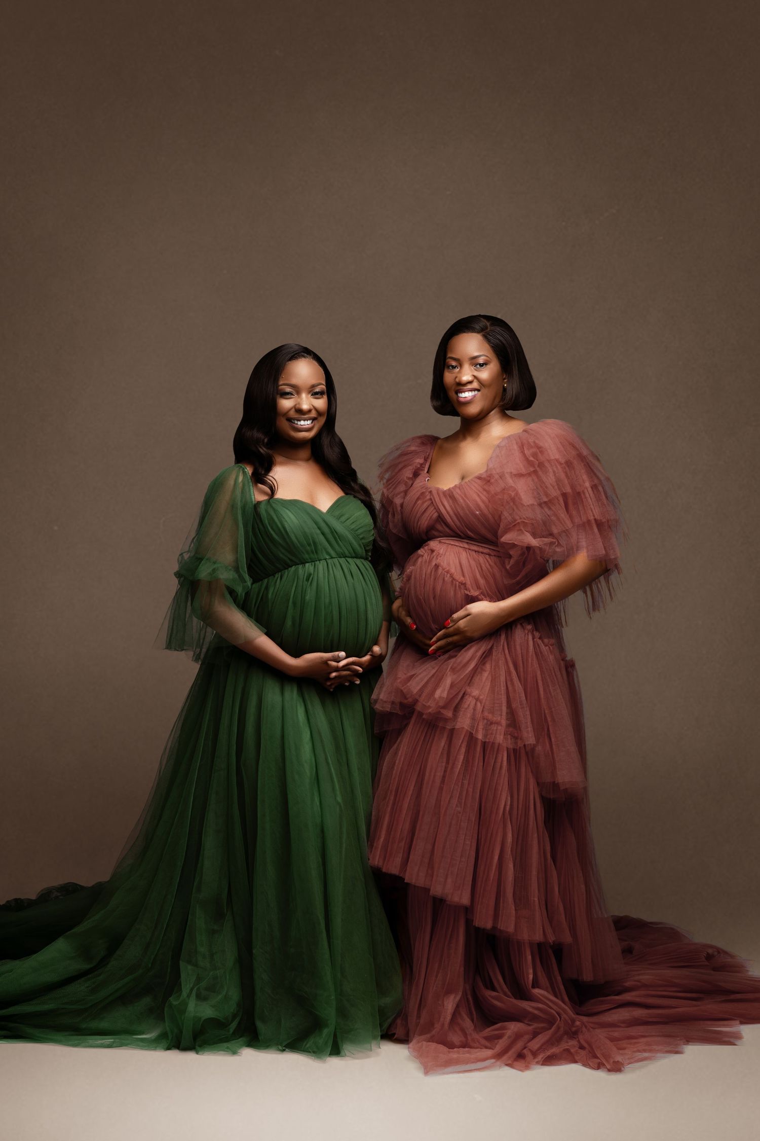 Two expectant mothers pose in elegant green and mauve tulle gowns against a brown backdrop.