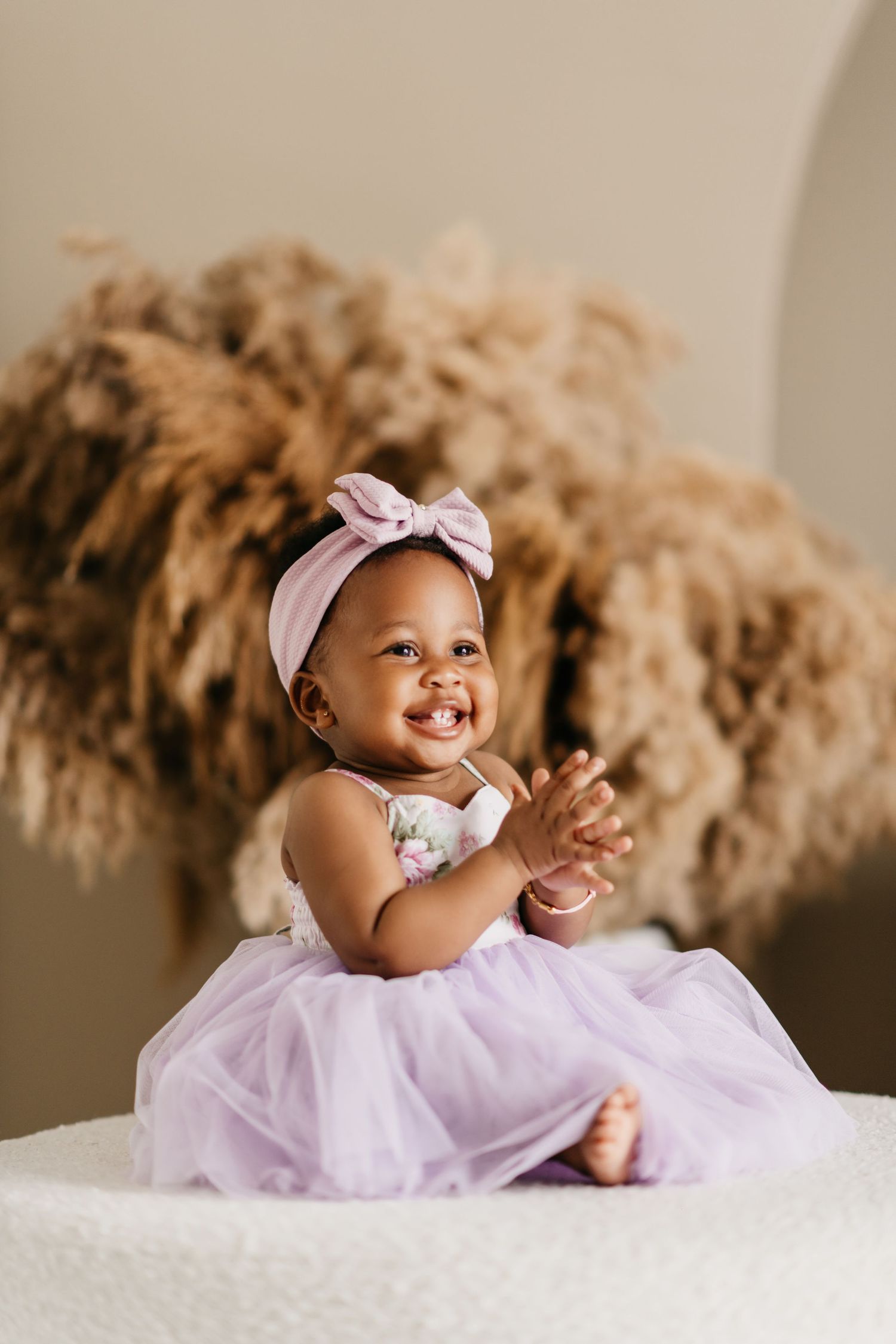 Baby in pink tulle dress and headband sits smiling in front of dried pampas grass.