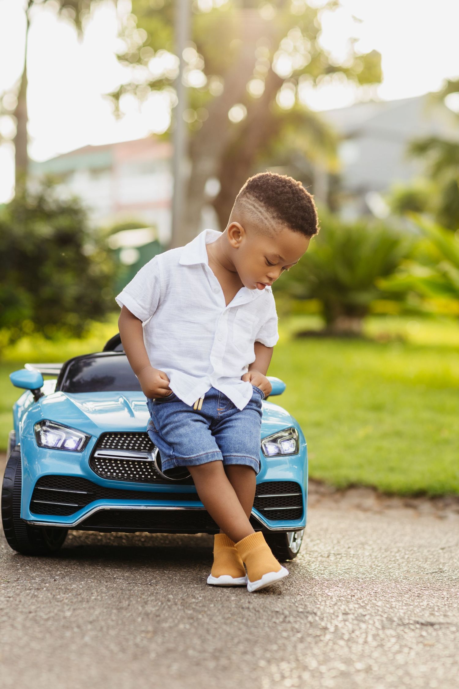 Young child sits on blue toy car outdoors wearing white shirt and denim shorts.