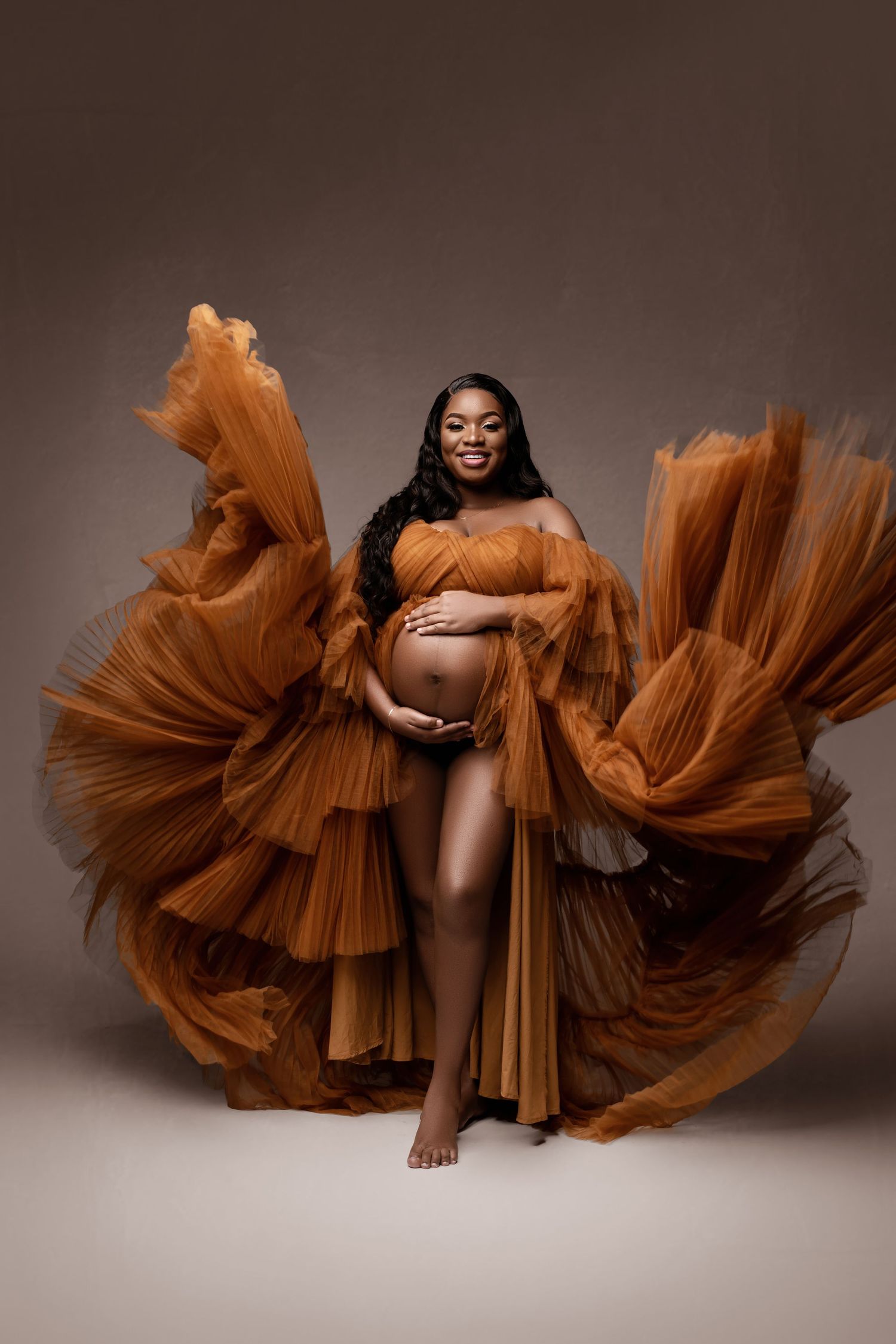 Maternity portrait featuring dramatic flowing rust-colored tulle fabric against brown background.