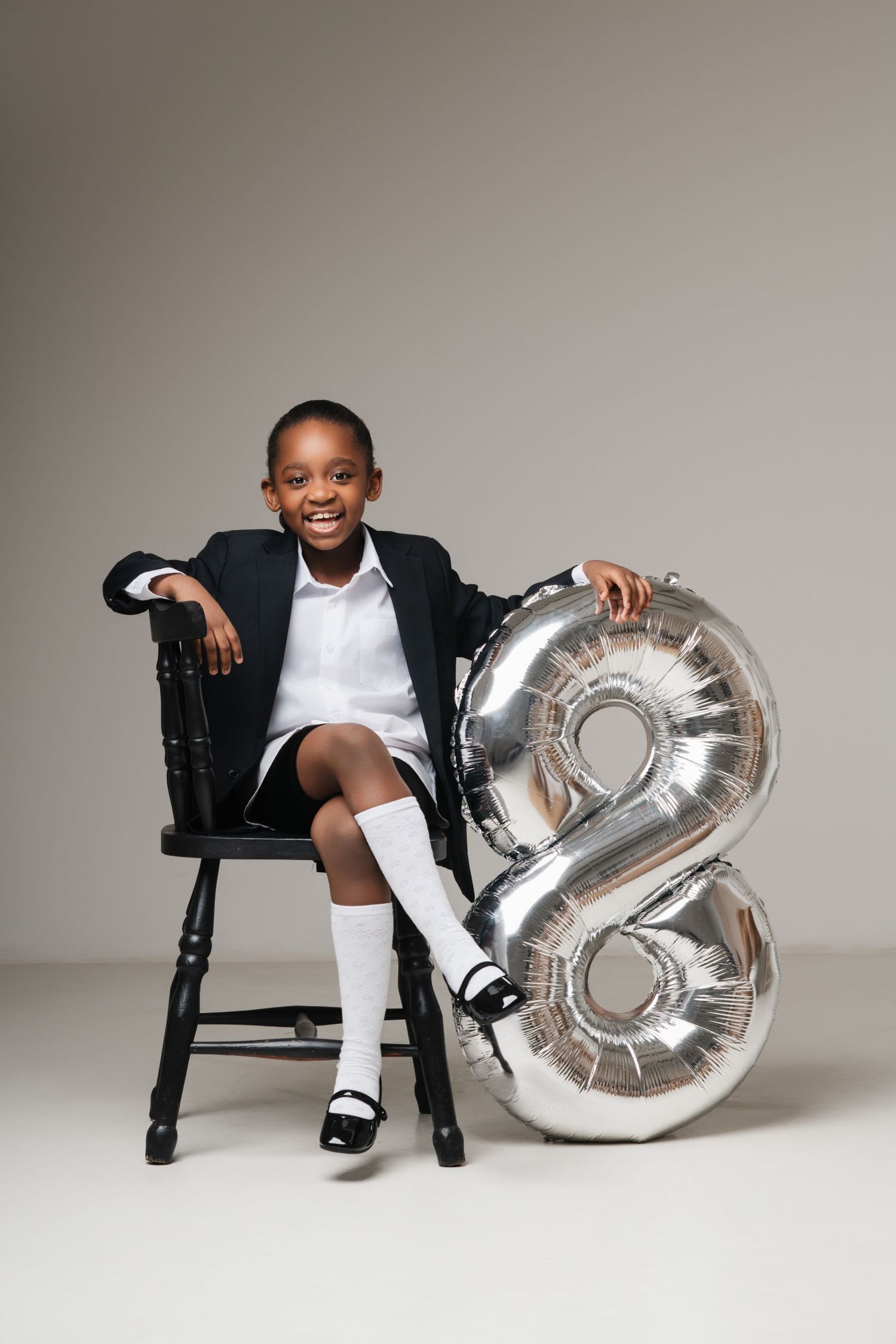 A child dressed in a black blazer and white shirt sits on a wooden chair next to a silver number 8 balloon.