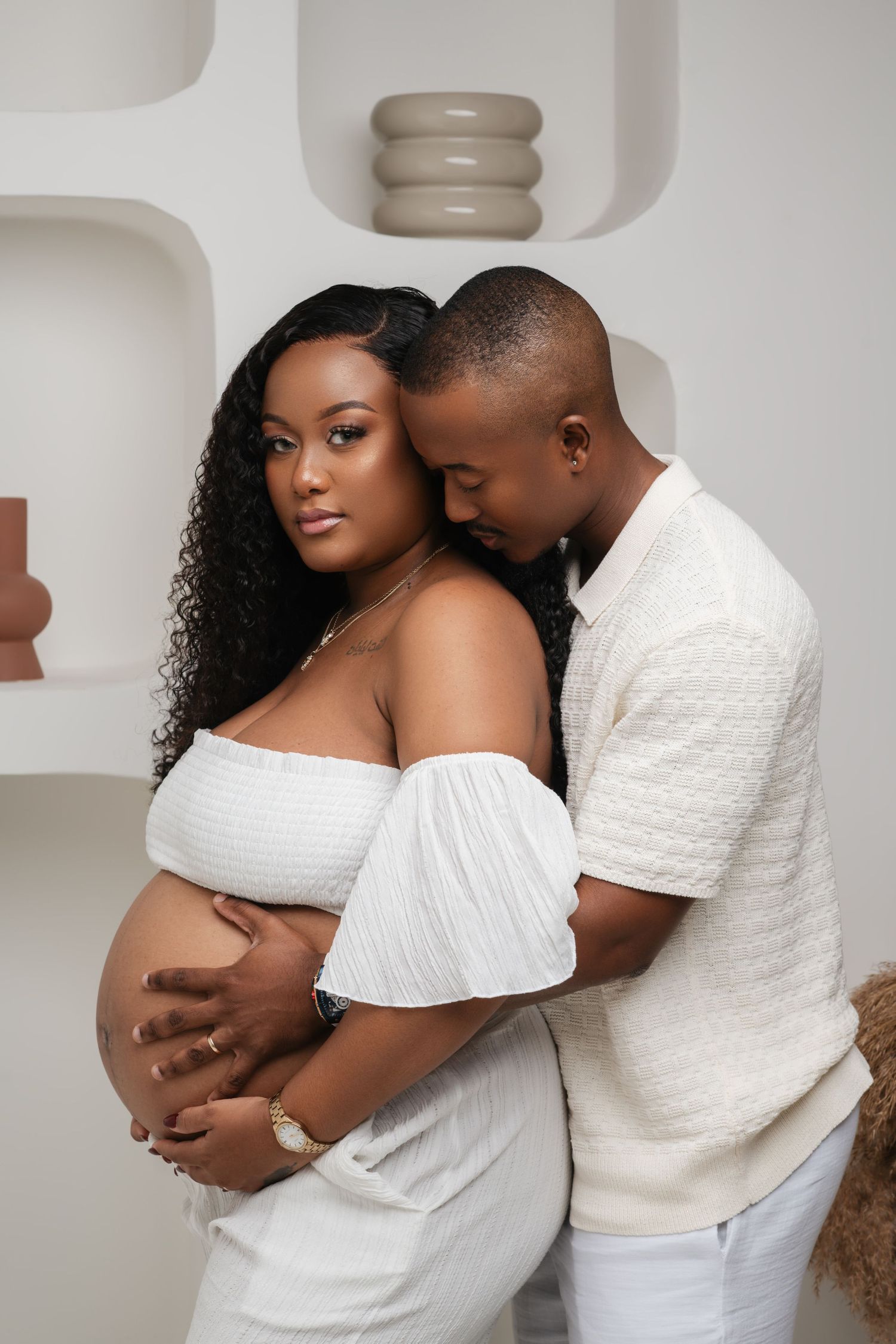 Couple in white outfits embrace during maternity photoshoot against neutral background.