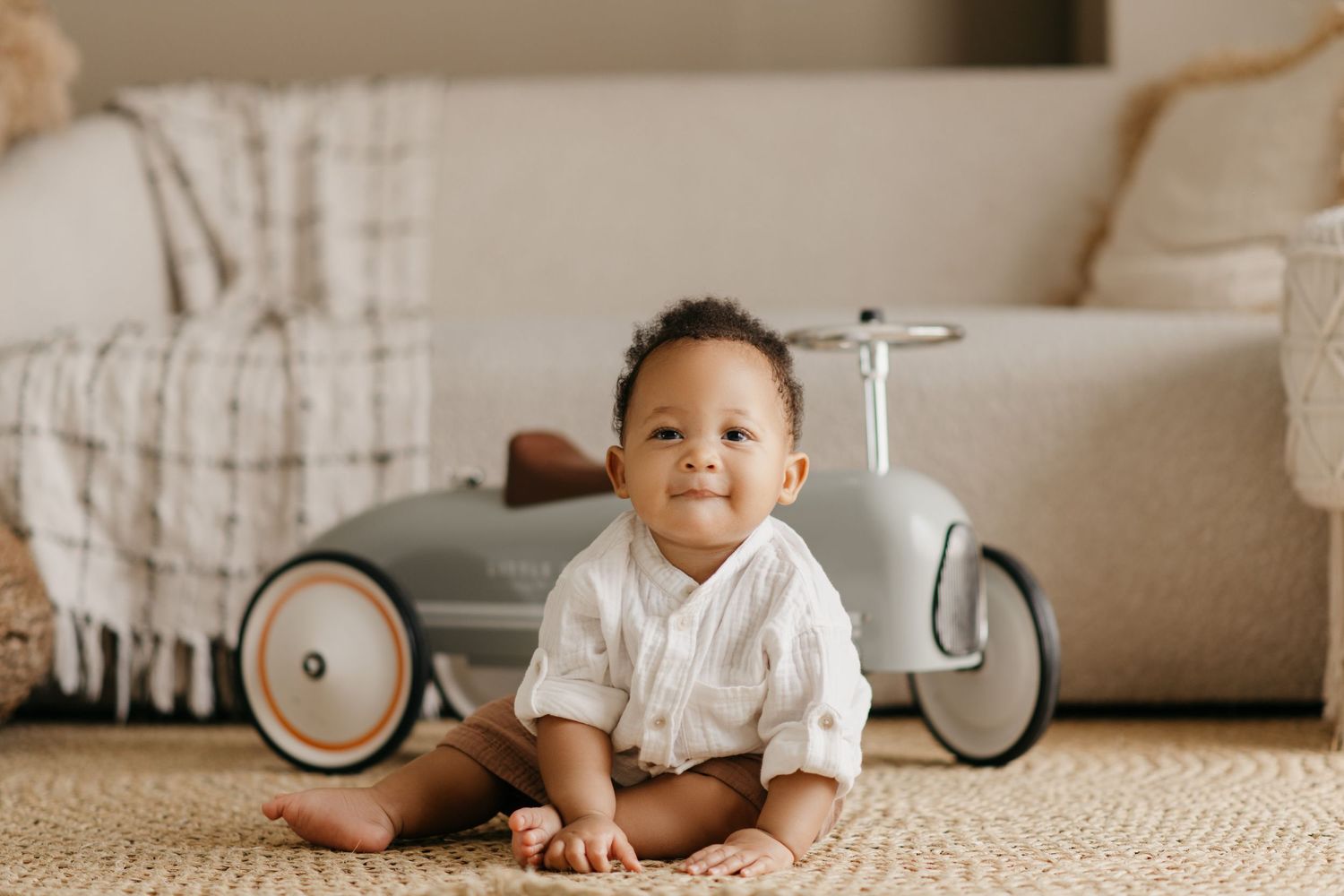 Baby in white shirt sits on carpet next to vintage-style toy ride-on car.