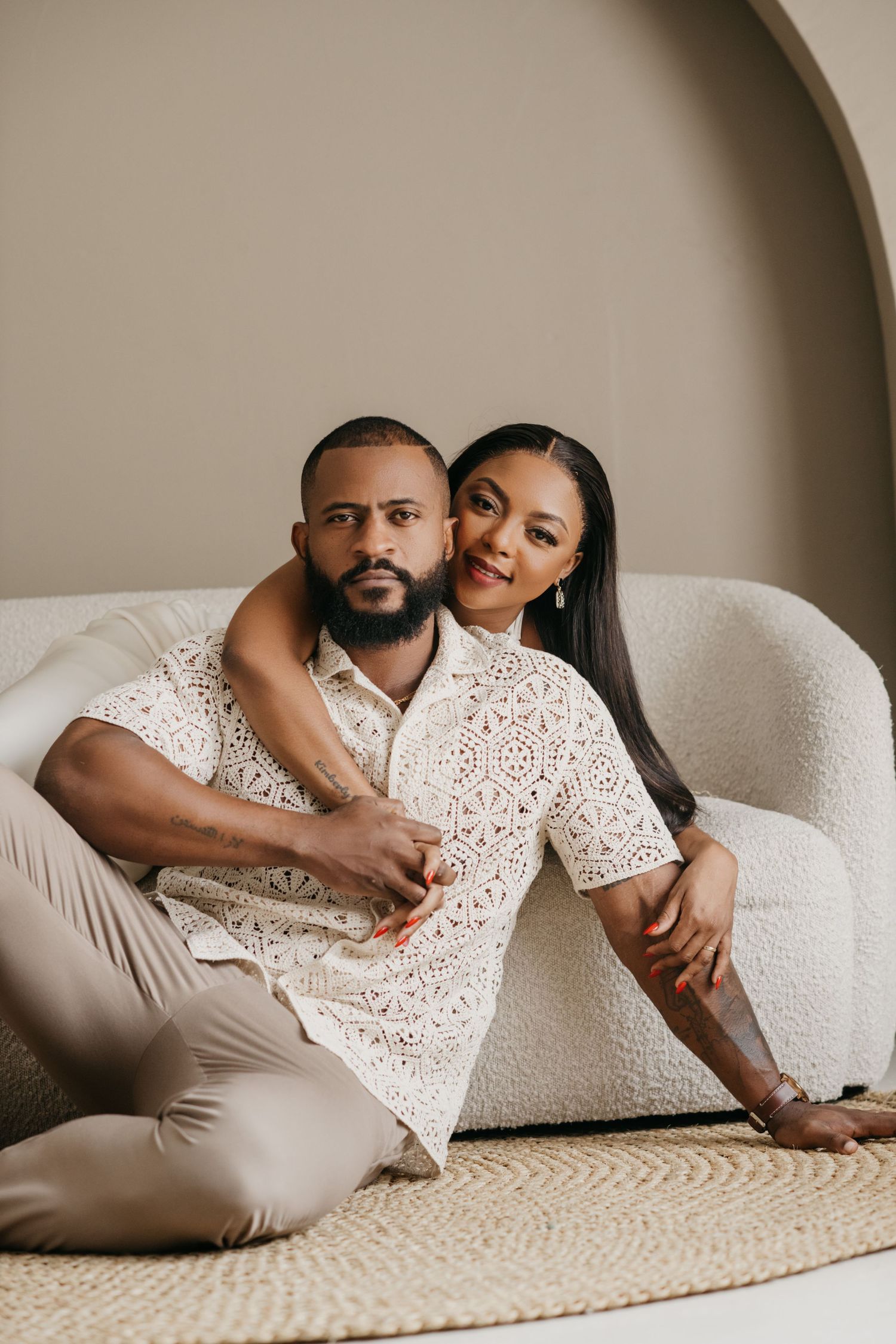 A couple in matching white lace outfits embraces while sitting on a plush chair against a neutral backdrop.