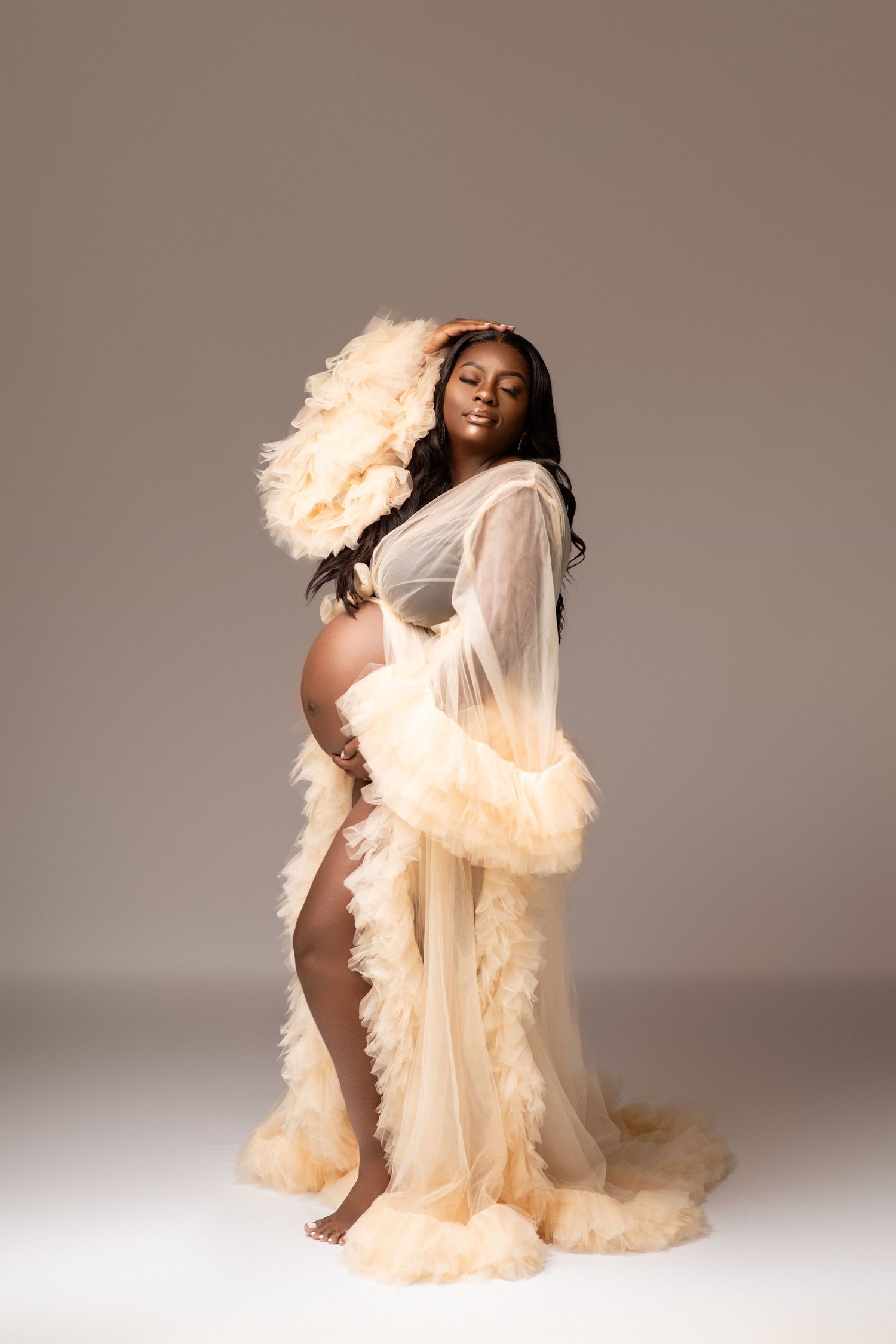 Maternity photo with flowing cream-colored sheer gown against grey background.