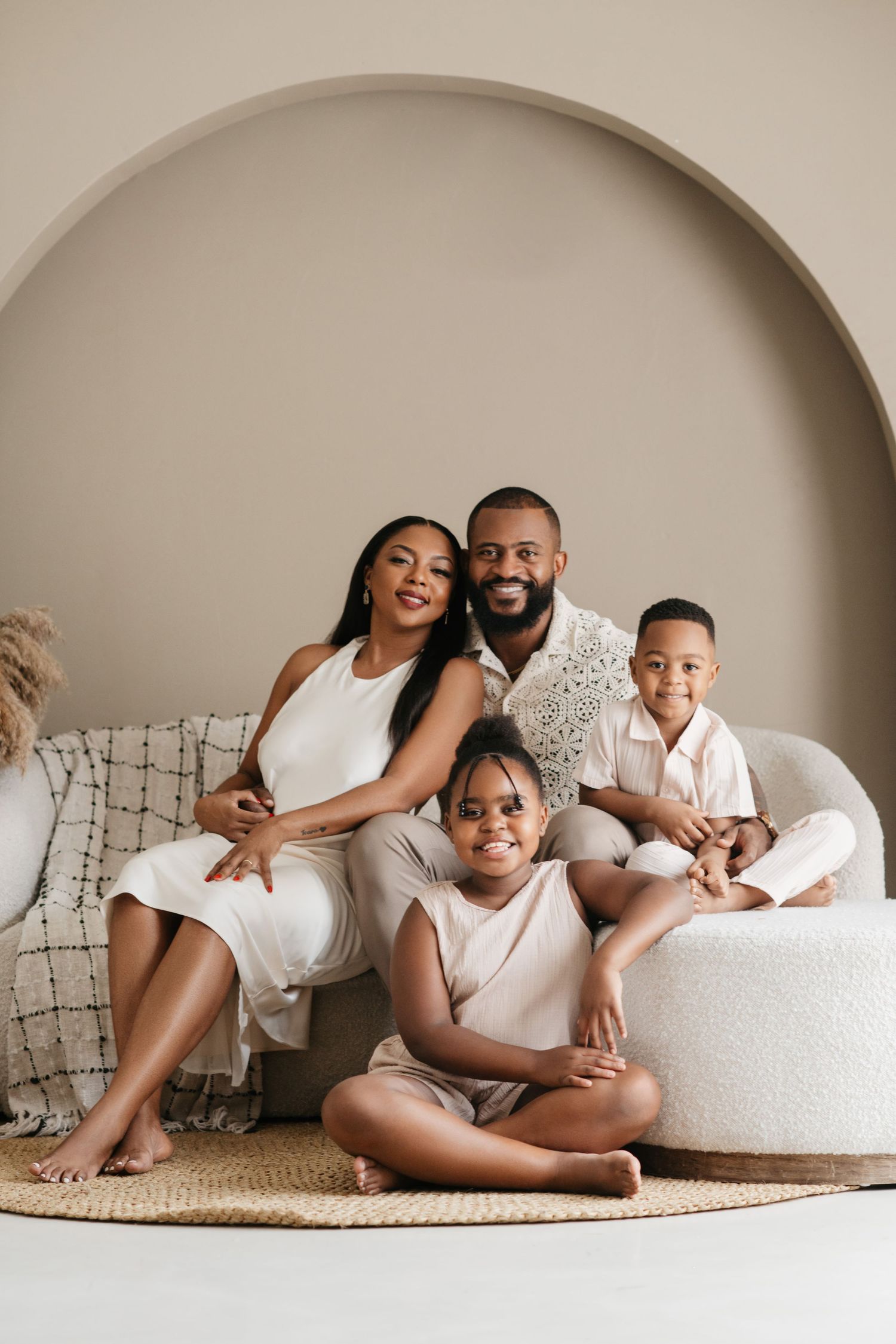 A family wearing coordinated white outfits sits together on a cream-colored chair against a neutral wall and circular arch.