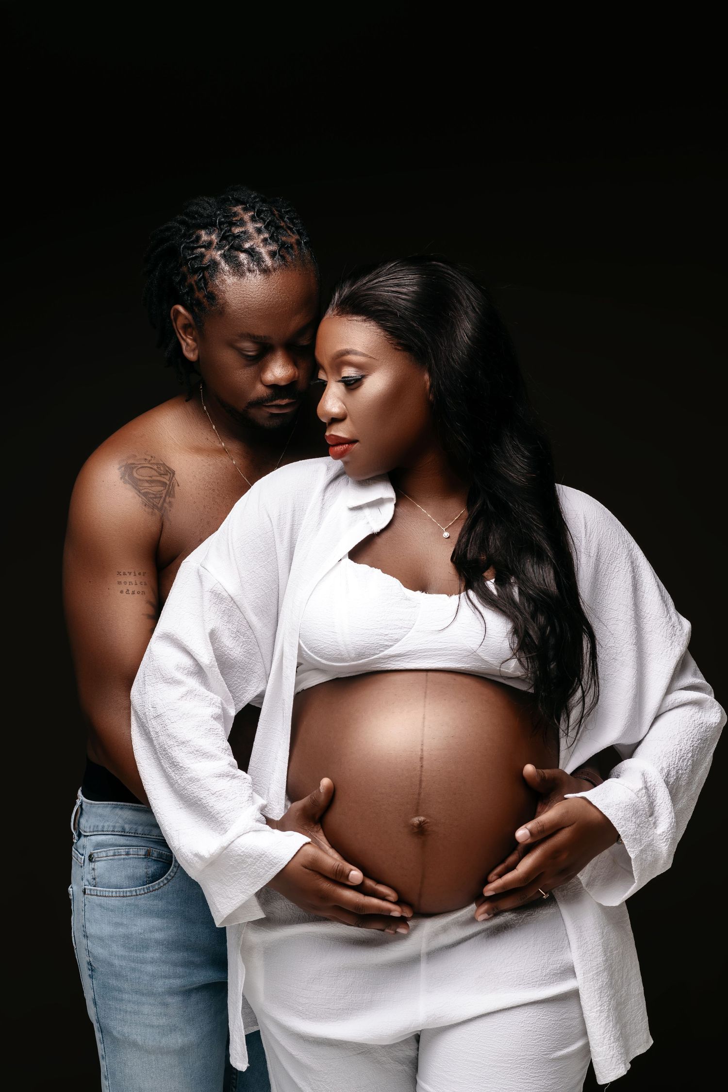 Intimate maternity portrait with couple in white and denim against dark background.