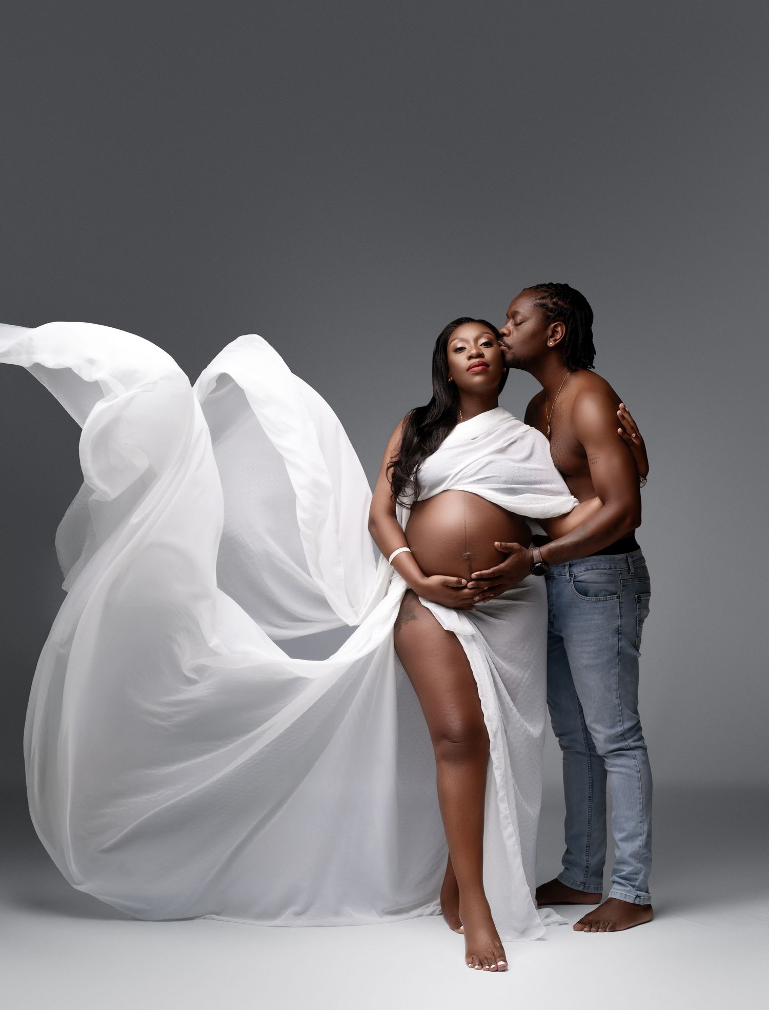 Artistic maternity photo with flowing white fabric against grey background showing intimate moment between couple.