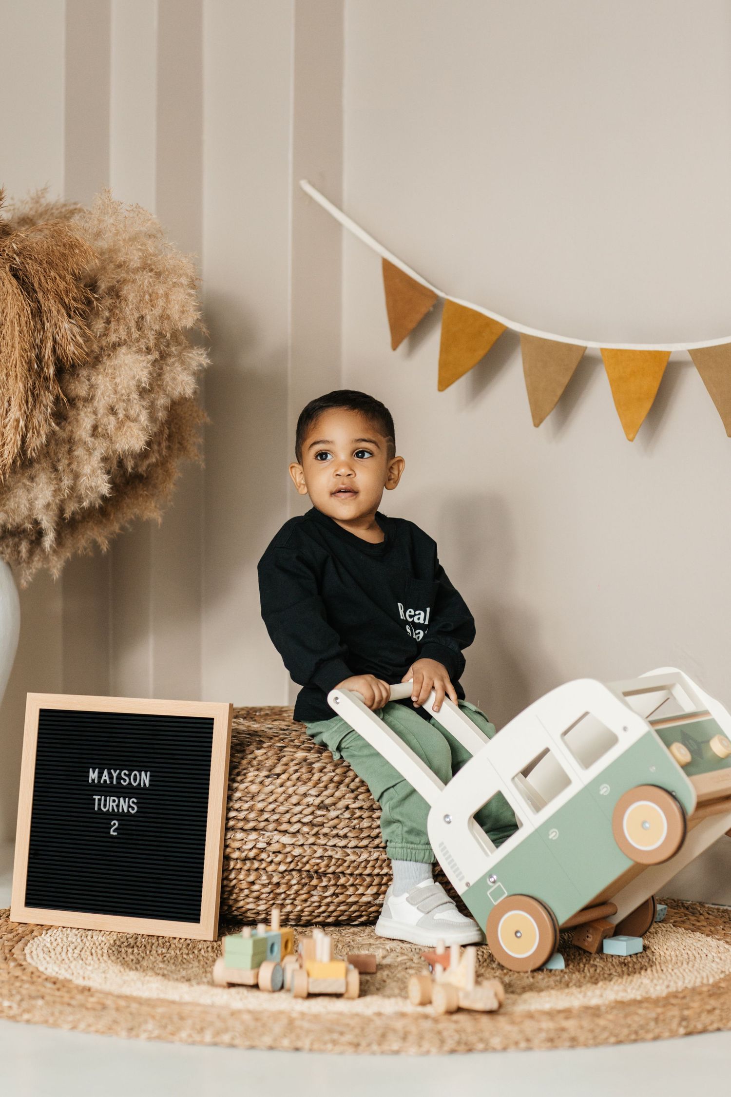 Child plays with wooden toy bus near decorative bunting and pampas grass.