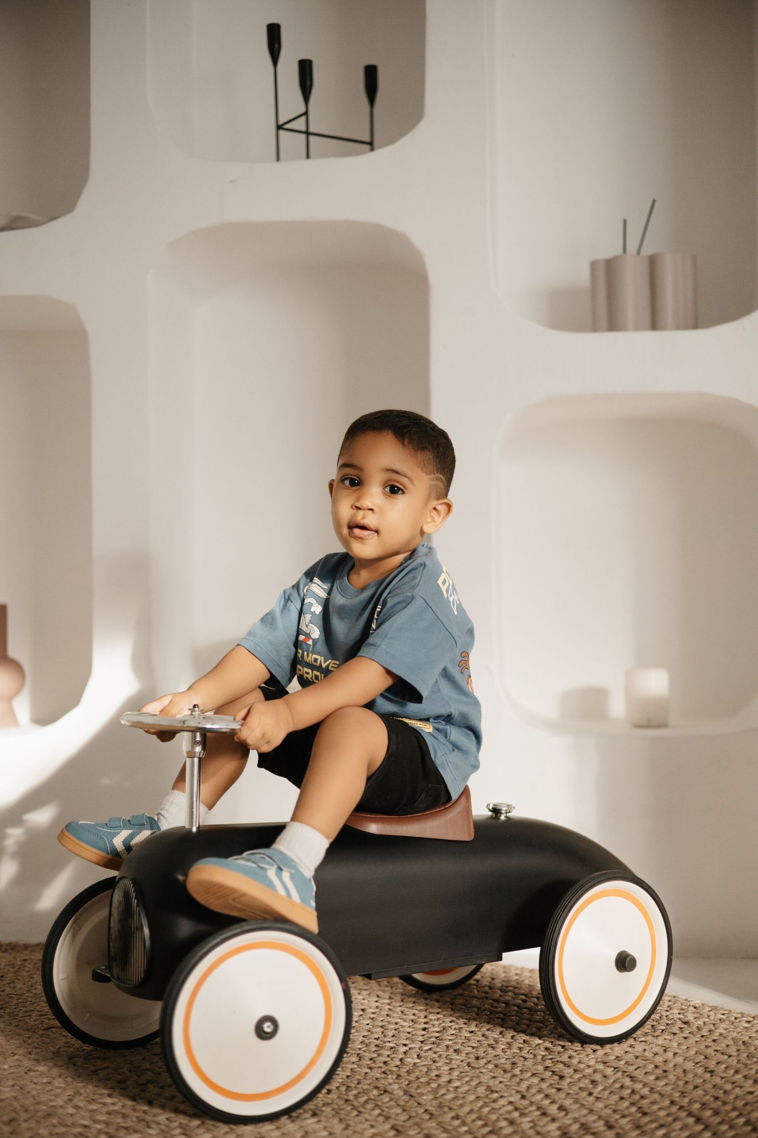Child rides vintage-style black pedal car on jute rug in minimalist room.