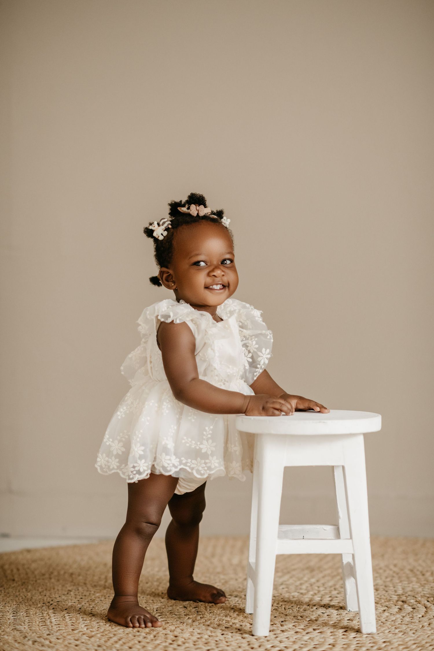Toddler in white lace dress stands smiling next to white stool on jute rug.