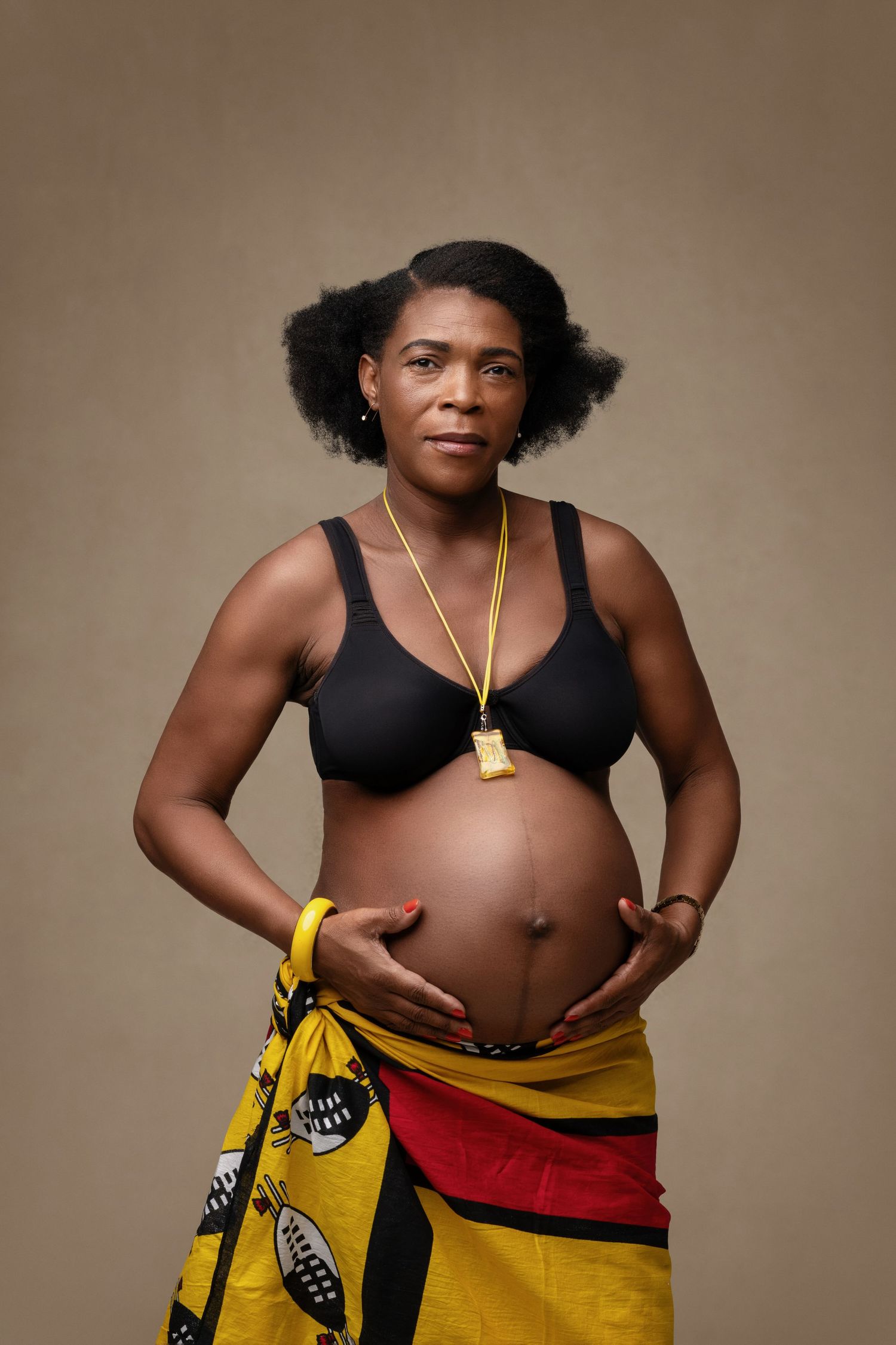 Expectant parent in traditional African wrap and black top poses against beige background.
