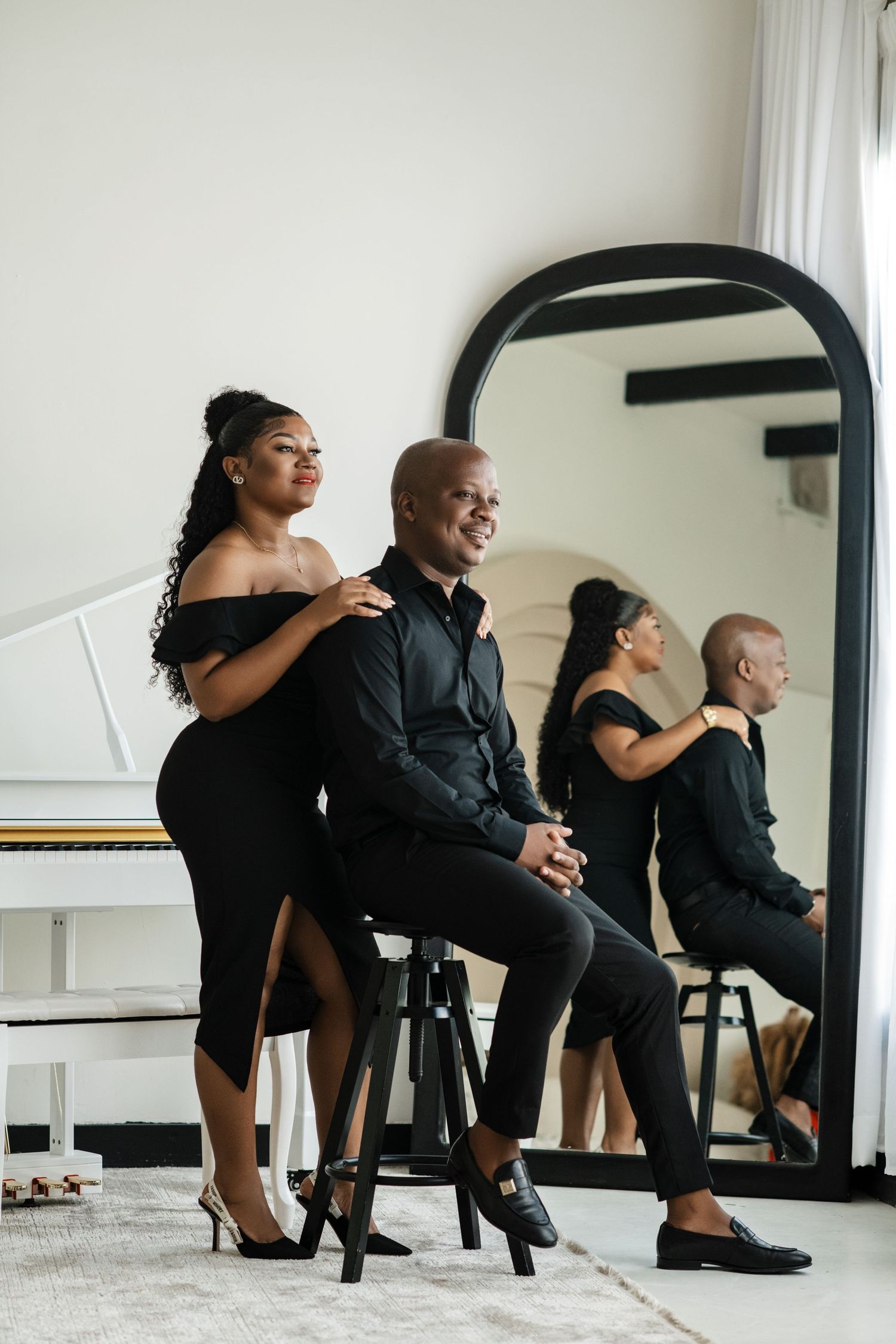 Elegant couple poses by white piano and mirror in stylish black outfits for artistic photoshoot.