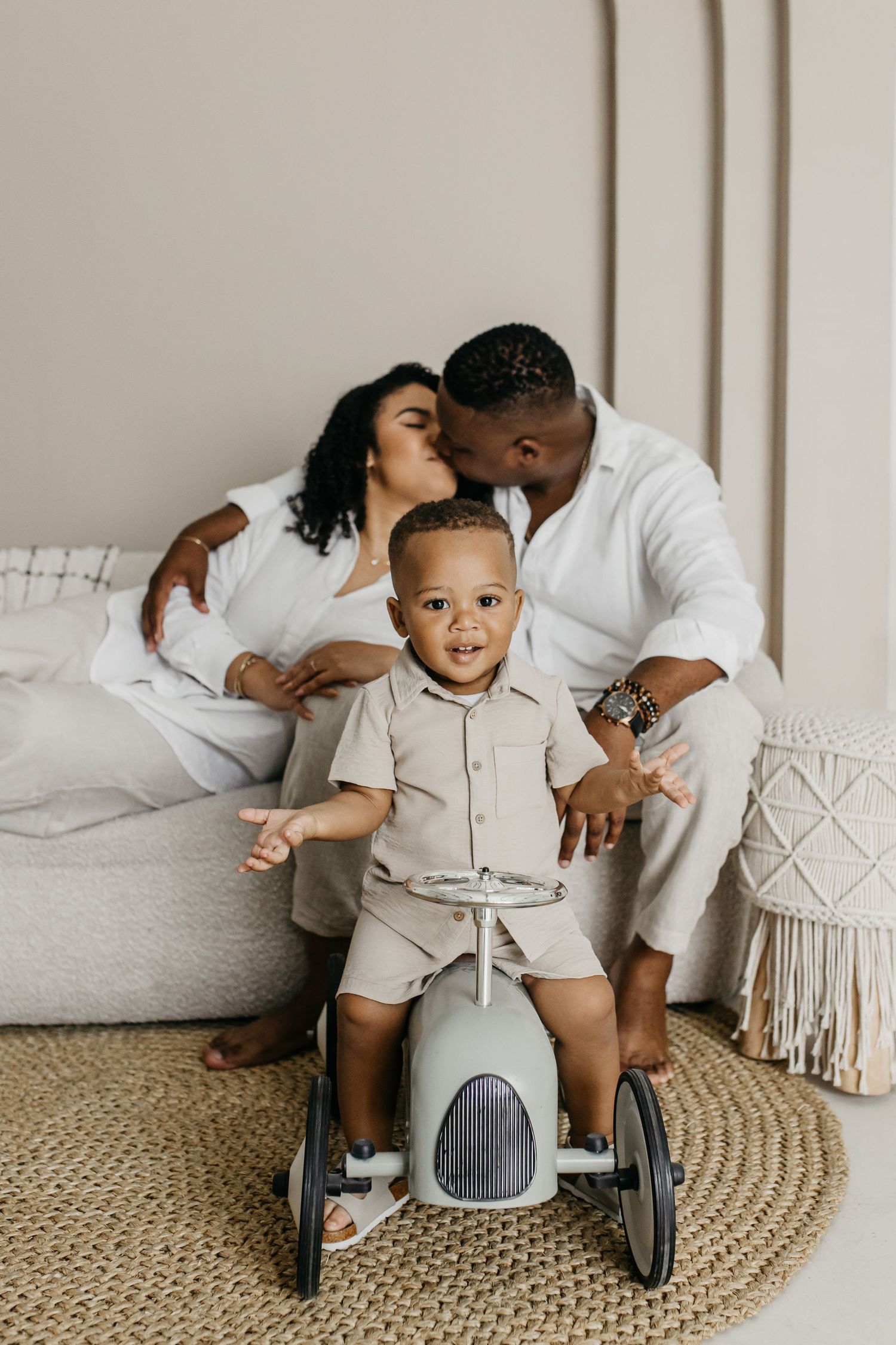 A toddler rides a vintage-style toy car while parents share a tender moment on the couch.