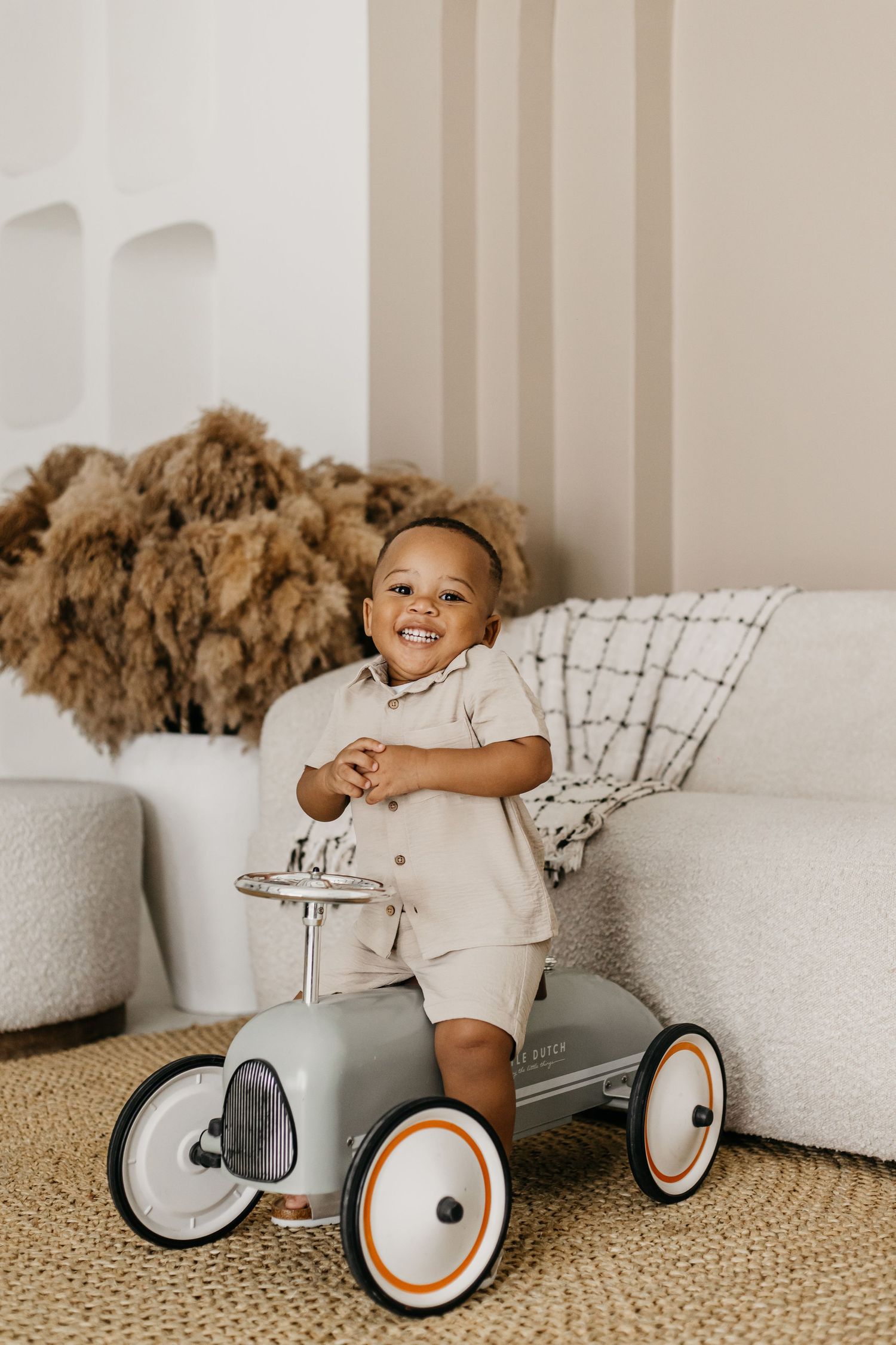Child sitting on a vintage-style gray metal ride-on toy car with white wheels on a beige carpet near white furniture.