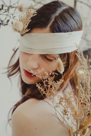 Artistic portrait with white blindfold and dried flowers creating ethereal mood in soft lighting.
