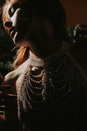 Dramatic portrait in moody lighting shows pearl necklace details in warm shadows against dark background.