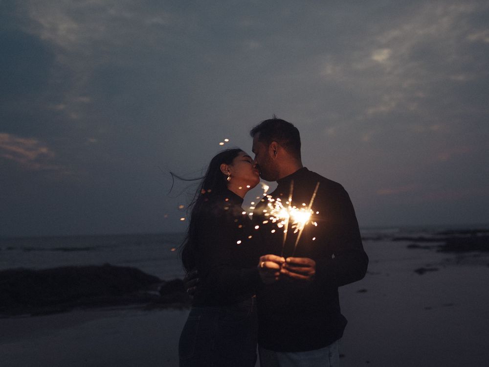 Couple holding sparklers on a beach at dusk, sharing a romantic moment under a cloudy evening sky.
