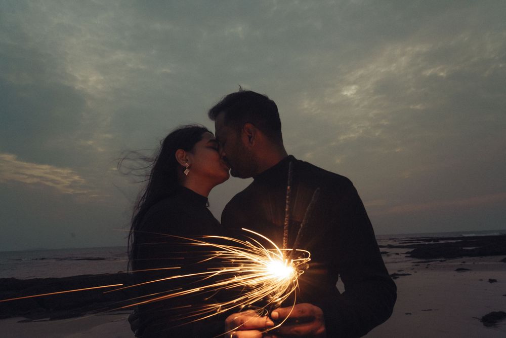 Romantic couple kissing on a beach at dusk while holding a sparkling sparkler, with dramatic cloudy sky behind them.
