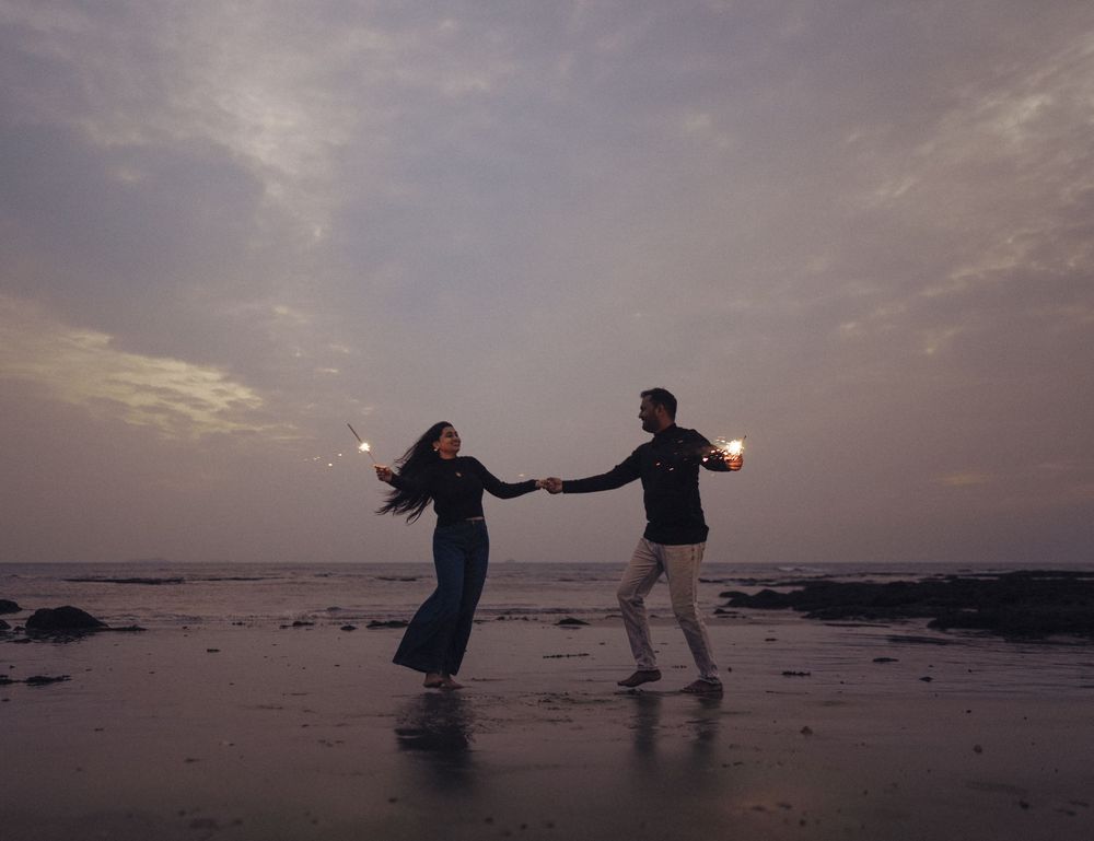 Couple holding sparklers dancing on a beach at dusk with cloudy sky backdrop.