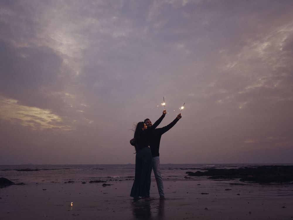 Couple holding sparklers on a beach at dusk, embracing against a dramatic cloudy sky at sunset.