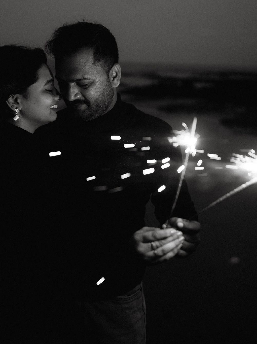 Black and white photo of a couple holding a sparkler together at night, sharing a romantic moment outdoors.