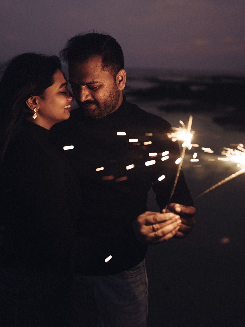 Couple in black outfits holding sparklers together at dusk, sharing a romantic moment outdoors at night.