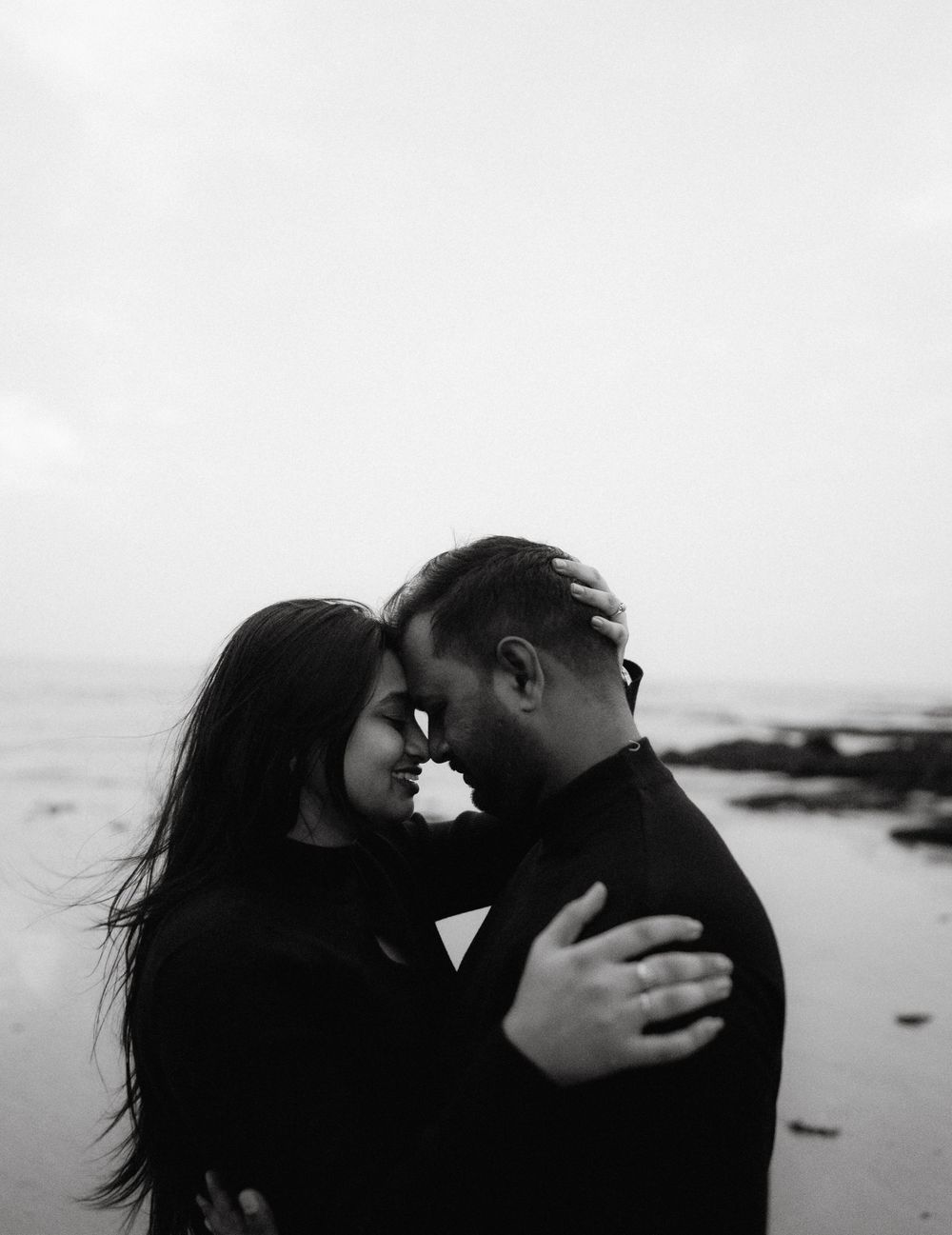 Black and white photo of a romantic couple embracing on a beach, touching foreheads tenderly.