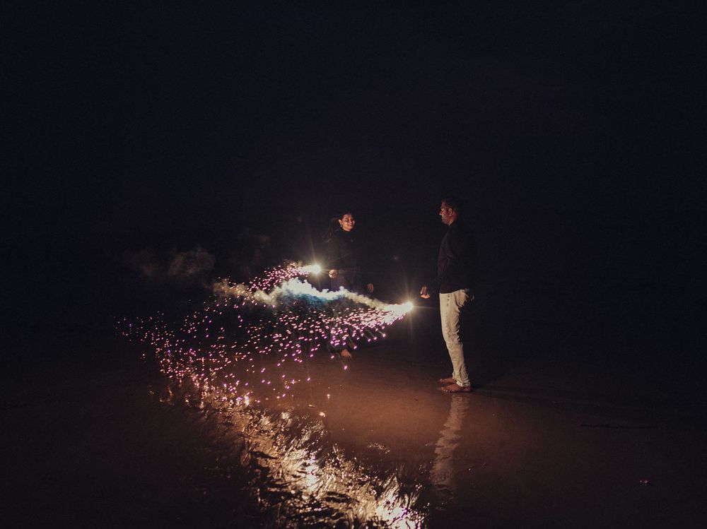 Two people lighting fireworks on a dark beach at night with colorful sparks illuminating the ground.