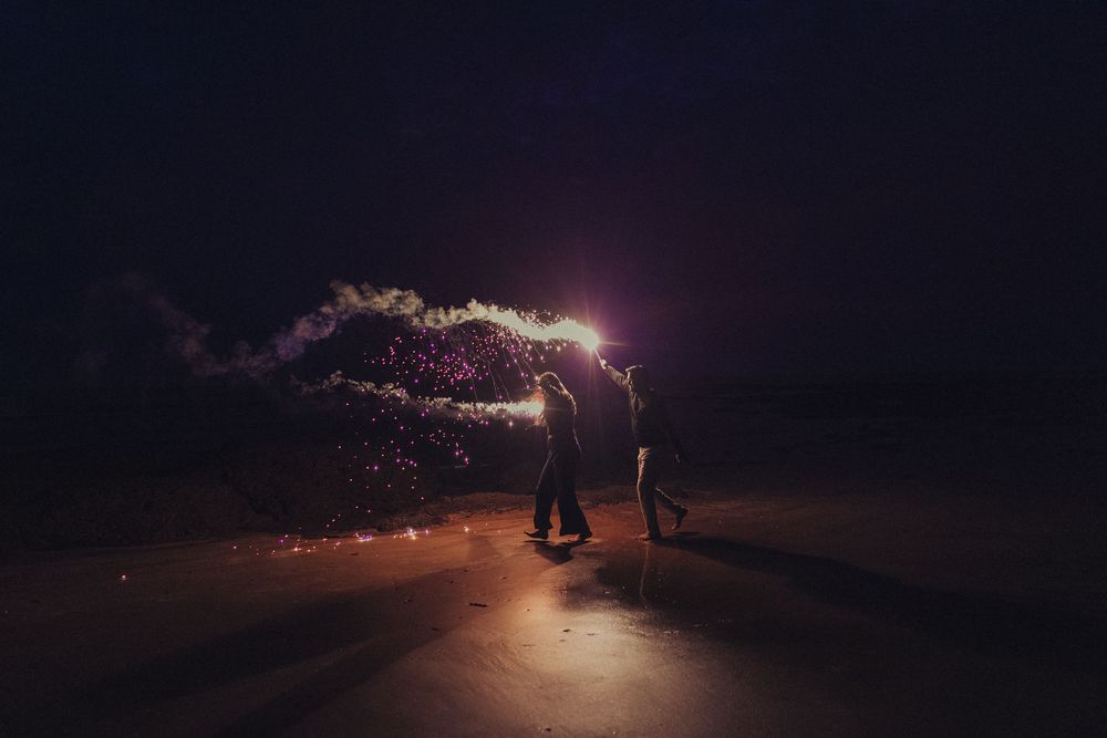 Two people holding sparklers at night on a road with colorful smoke and sparks illuminating the dark sky.