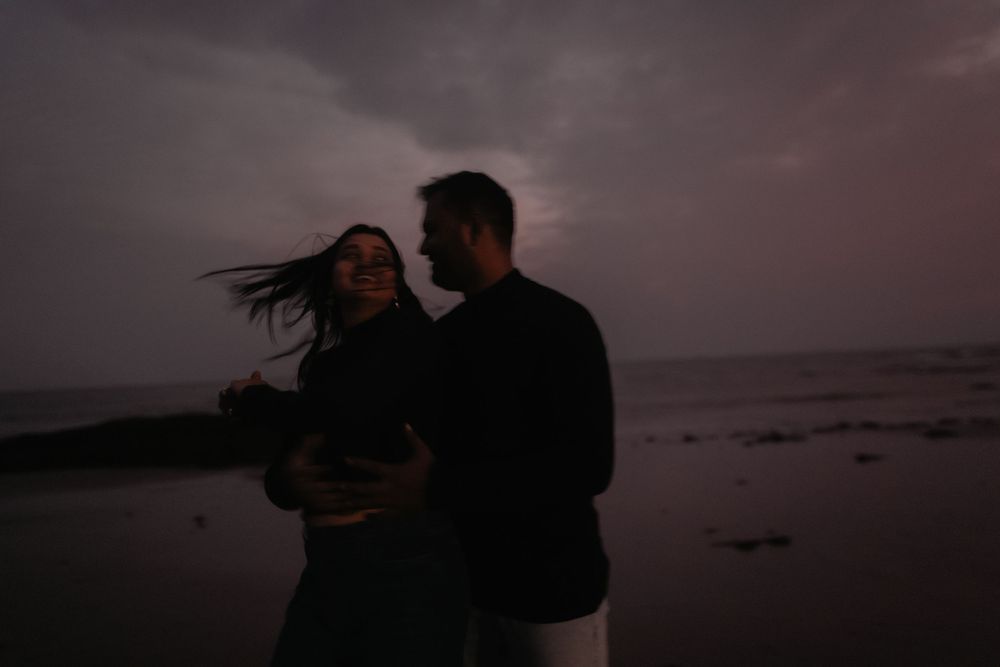 Romantic couple embracing on a beach at dusk with dramatic cloudy sky and ocean in the background.