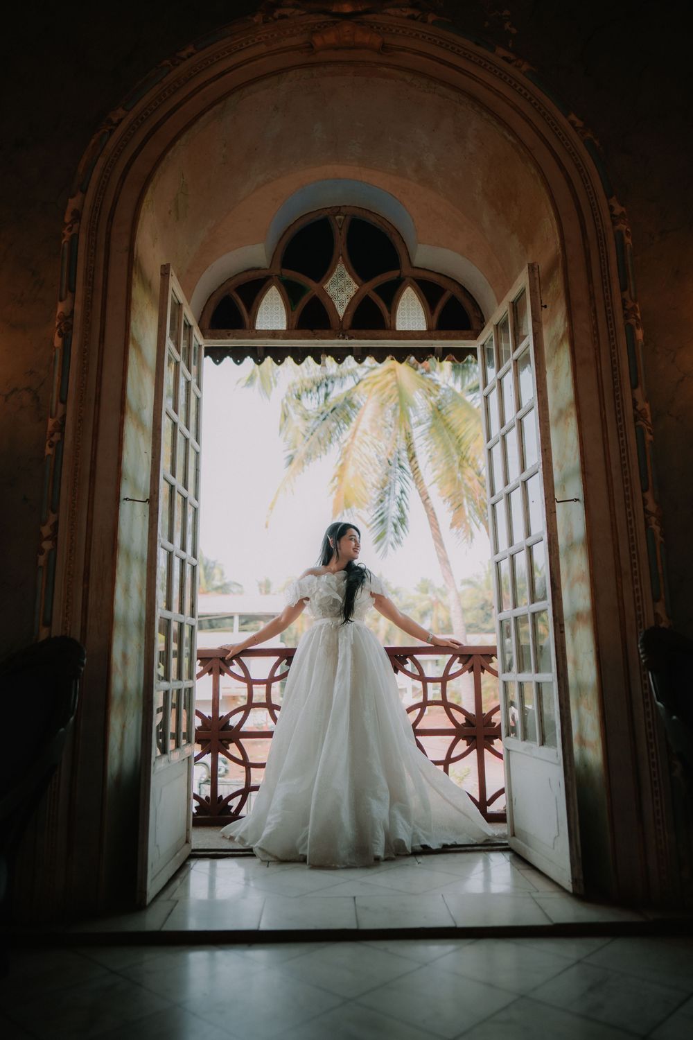 Bride in white ballgown standing in ornate arched doorway with palm tree visible on outdoor balcony.