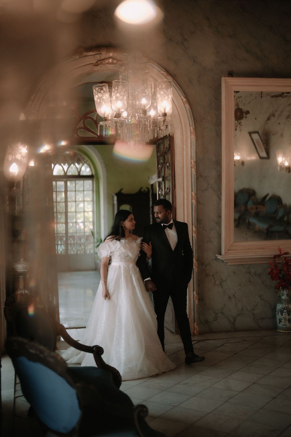Bride in white gown and groom in black tuxedo share a romantic moment in an elegant chandelier-lit venue.