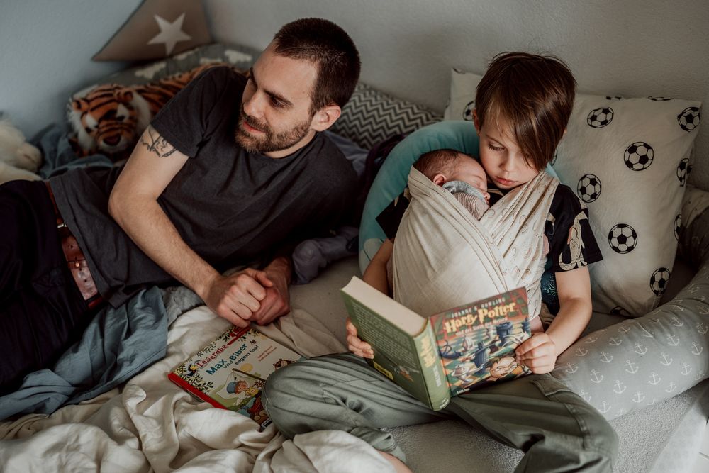 Grosse Schwester hat ihren kleinen Bruder im Tragetuch und liest ein Buch neben ihrem Papa