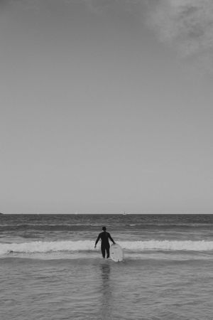 A silhouetted figure wades into ocean waves under an overcast sky in black and white.