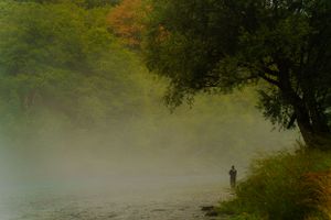 Silhouette of person fishing by misty river surrounded by autumn trees.