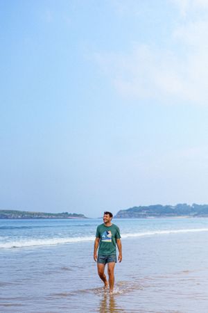 A beachgoer walks along the shoreline on a sunny day with coastal cliffs in the distance.