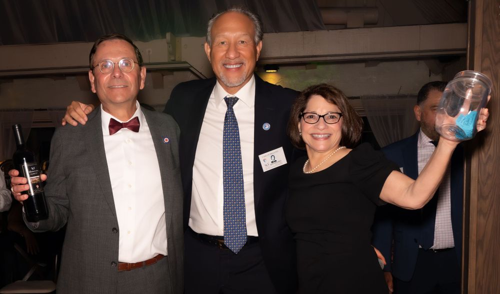 Three people at a formal event pose together, with a person in a bow tie, one in a business suit, and one wearing glasses.