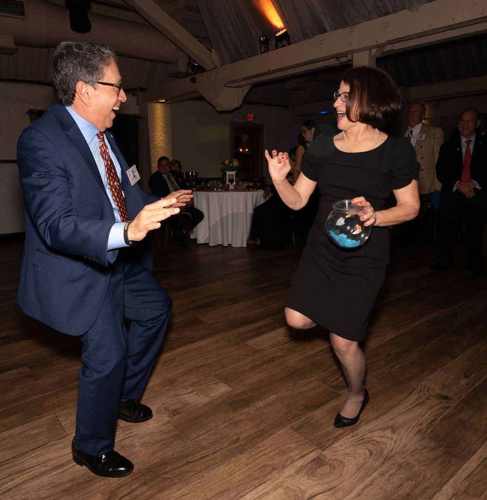 People dancing and having fun at an indoor event with wooden floors and white tablecloths in the background.
