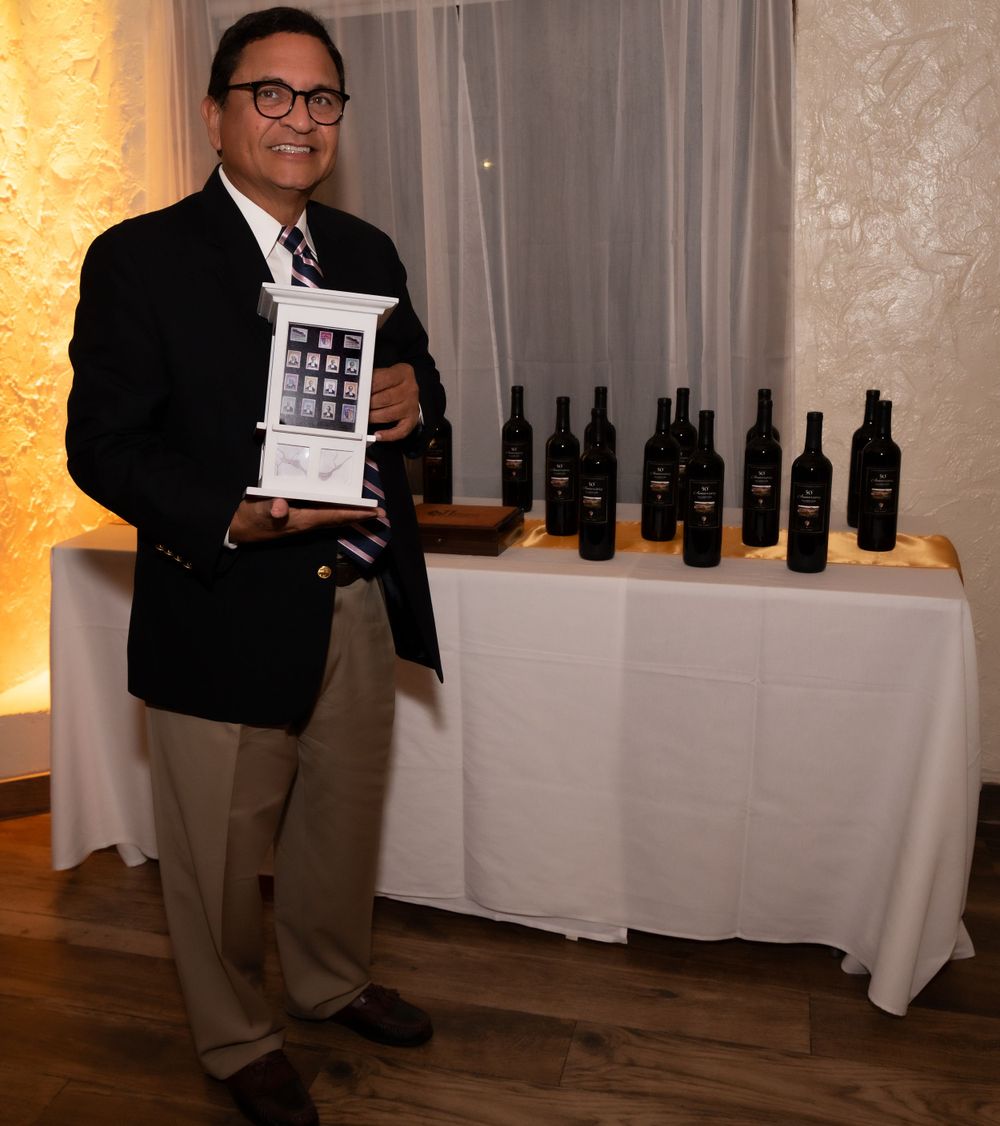 A presenter stands next to a table displaying wine bottles while holding a framed image in an elegant indoor setting.