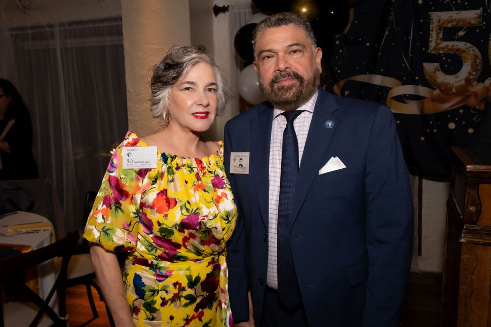 Two guests celebrate at a formal event, with one wearing a vibrant yellow floral dress and the other in a navy suit.
