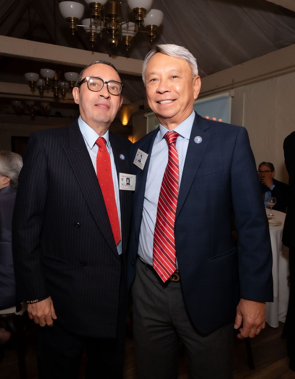 Two people in business suits and red ties stand together at an indoor event with chandeliers visible above.