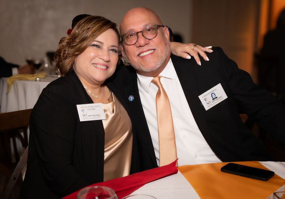 Two attendees wearing name tags share a warm moment at a formal event with a red tablecloth visible.