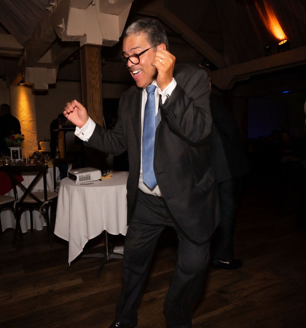 A person in a dark suit and blue tie dances energetically on a wooden floor at an event with white tablecloths.