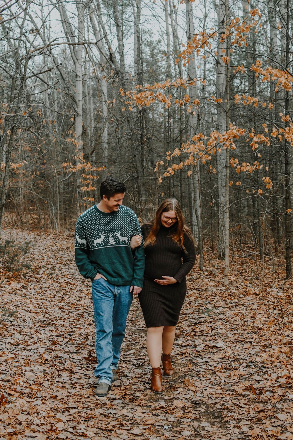 A couple walks together on a leaf-covered forest path during autumn, with orange foliage visible in the background.