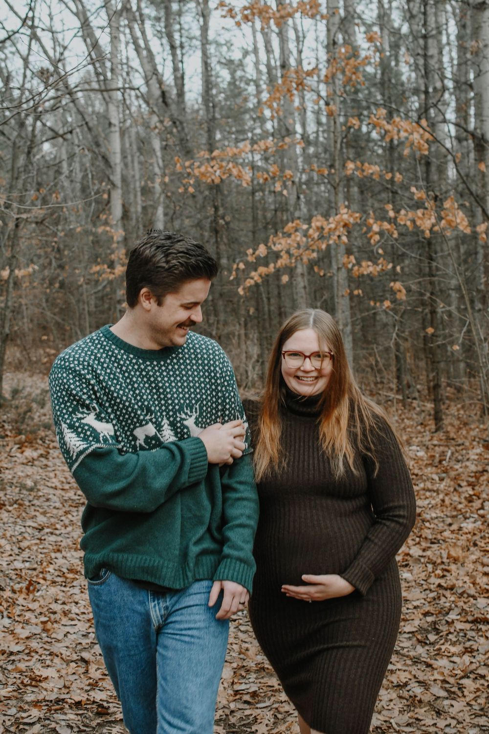 A couple in cozy winter sweaters walking together through a forest with autumn leaves on the ground.