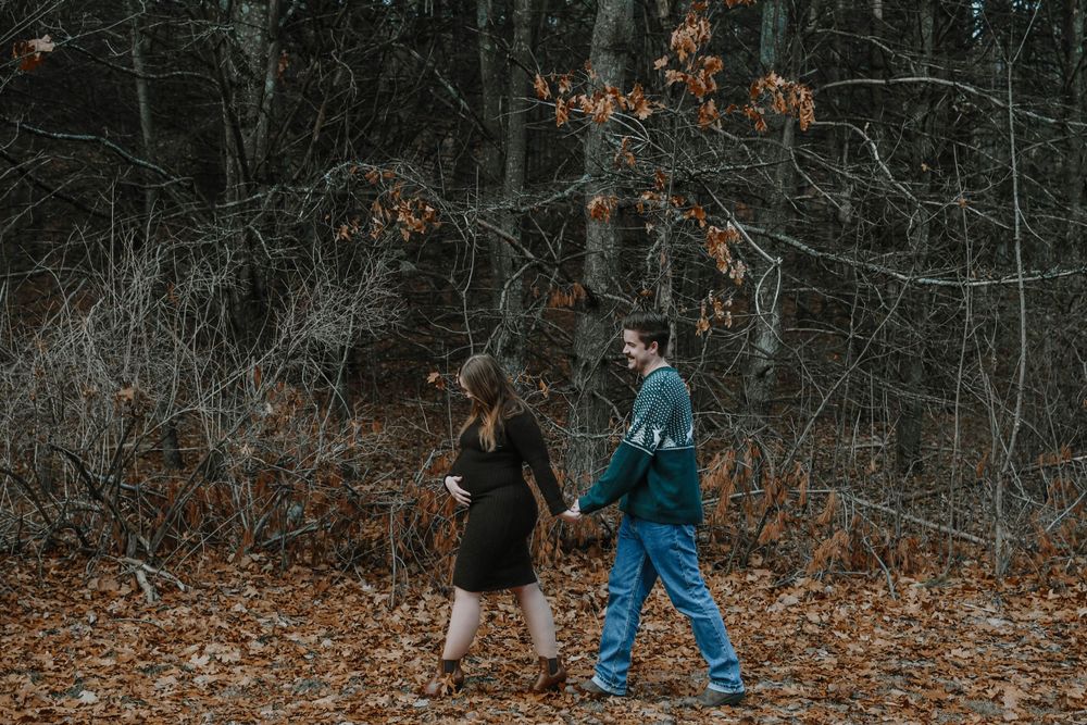 Two people walk through an autumn forest with fallen orange leaves and bare trees in moody lighting.
